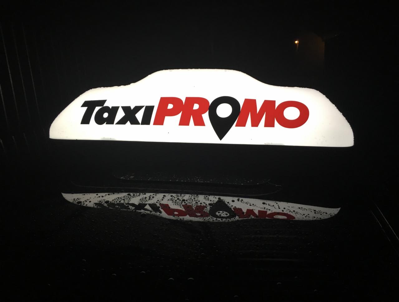 Podświetlony szyld Taxi PROMO z czarną ikoną lokalizacji zamiast litery 'O', widoczny na dachu taksówki, odbicie na mokrej karoserii.