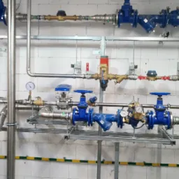 Kompleksowy system rur i zaworów z manometrami, zainstalowany na metalowej konstrukcji przy ścianie z białych pustaków, z widocznymi elementami instalacji wodnej i grzewczej.