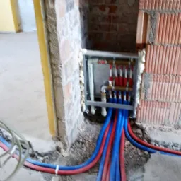 Rozdzielacz ogrzewania podłogowego w trakcie montażu, widoczne czerwone i niebieskie rury w bruzdach w ścianie, pianka montażowa uszczelniająca obudowę rozdzielacza.