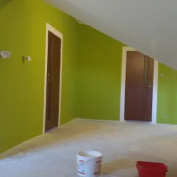 Świeżo pomalowane na zielono poddasze z dwiema brązowymi ościeżnicami drzwiowymi, widoczny kubeł z farbą i kuweta malarska na podłodze, ślady po taśmie malarskiej na ścianie.