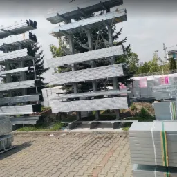 Magazyn zewnętrzny z metalowymi profilami i płytami budowlanymi na półkach regałów, widoczne także materiały izolacyjne w tle.