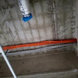 Kamil Srokosz ELEKTRO-KAM - Nierówny betonowy sufit z przymocowanymi wiązkami pomarańczowych i szarych kabli elektrycznych, widoczna instalacja wentylacyjna z niebieskim zabezpieczeniem.