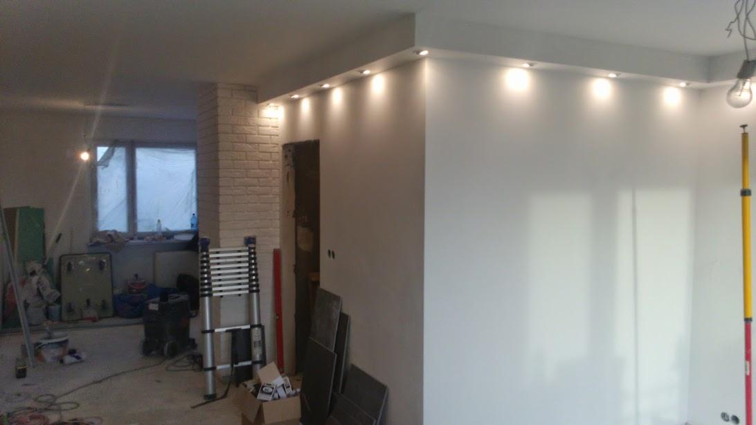 Wnętrze w trakcie remontu z zamontowanymi punktowymi źródłami światła w suficie podwieszanym, widoczna drabina, narzędzia i zabezpieczone okno folią malarską.