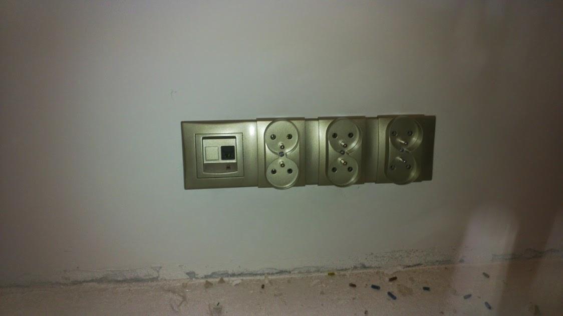Rząd gniazdek elektrycznych i włącznik w złotej ramce zamontowany na ścianie, widoczne ślady po remoncie na dole ściany.