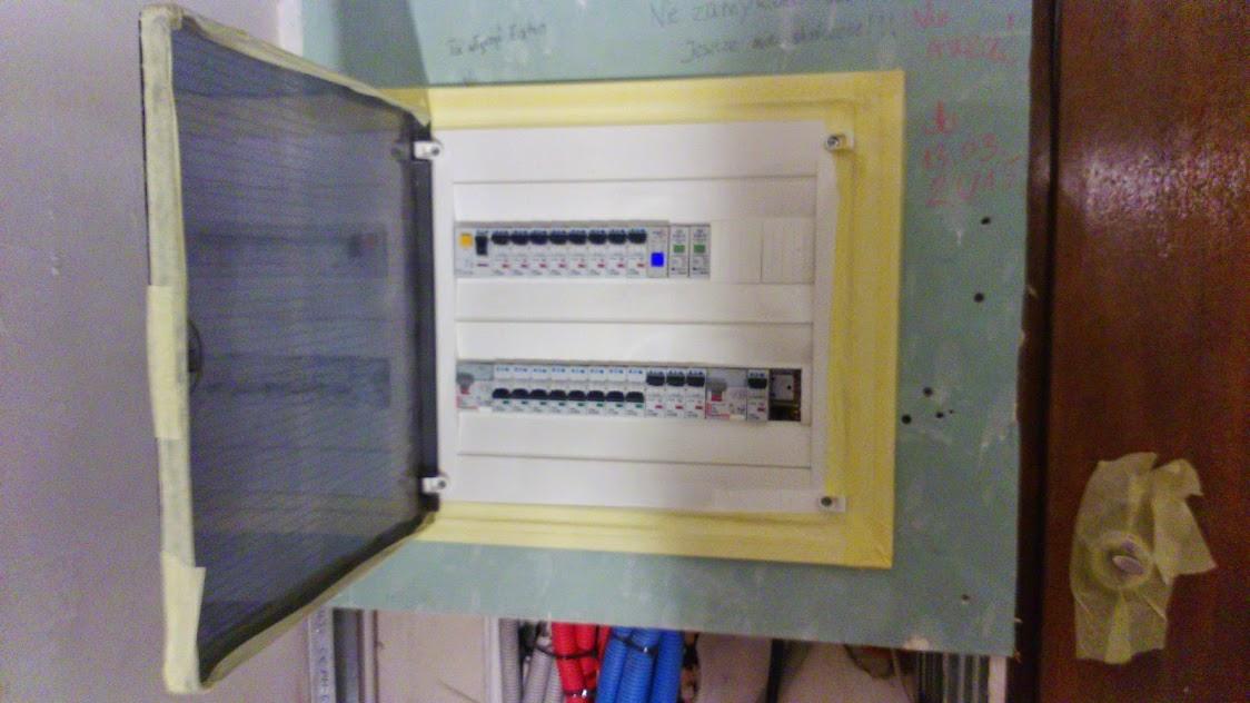 Otwarta skrzynka bezpiecznikowa z widocznymi bezpiecznikami, zamontowana na ścianie, z widocznymi przewodami w dolnej części kadru. Ściana w stanie surowym z widocznymi notatkami.