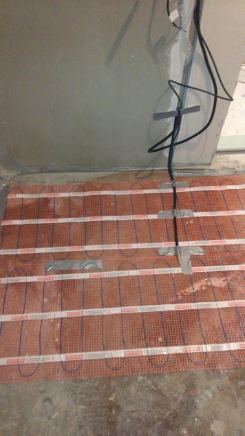 Mata grzewcza podłogowa z przewodami elektrycznymi, częściowo zamocowana taśmą, na tle surowej ściany i podłogi.