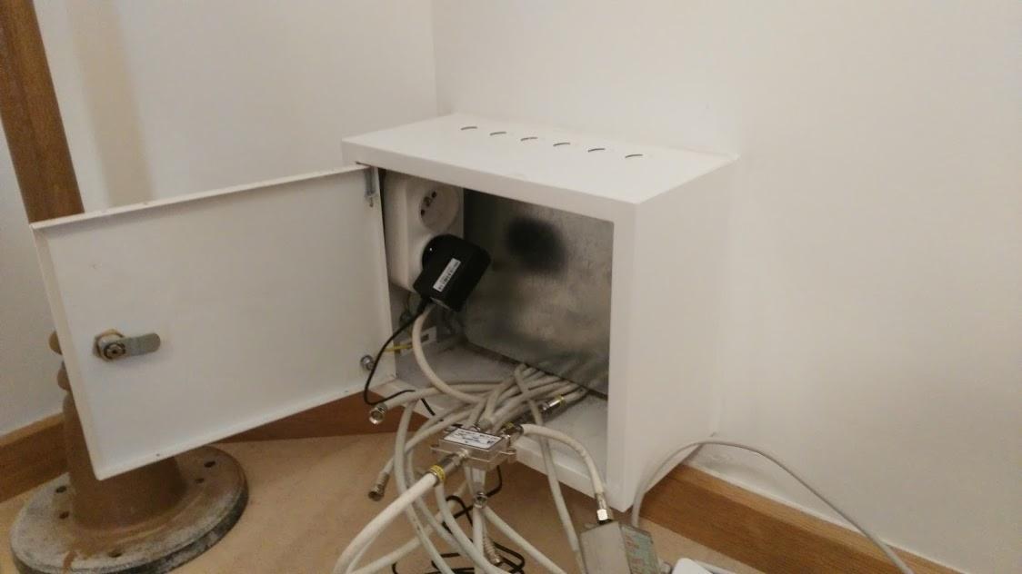 Otwarta, biała skrzynka z widocznym gniazdkiem elektrycznym, wzmacniaczem sygnału i plątaniną kabli koncentrycznych.