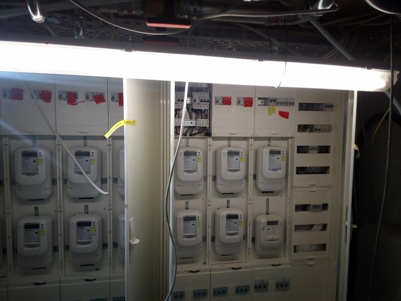 Rozdzielnica elektryczna z zamontowanymi licznikami energii elektrycznej, widoczne przewody i zabezpieczenia, oświetlenie jarzeniowe u góry.