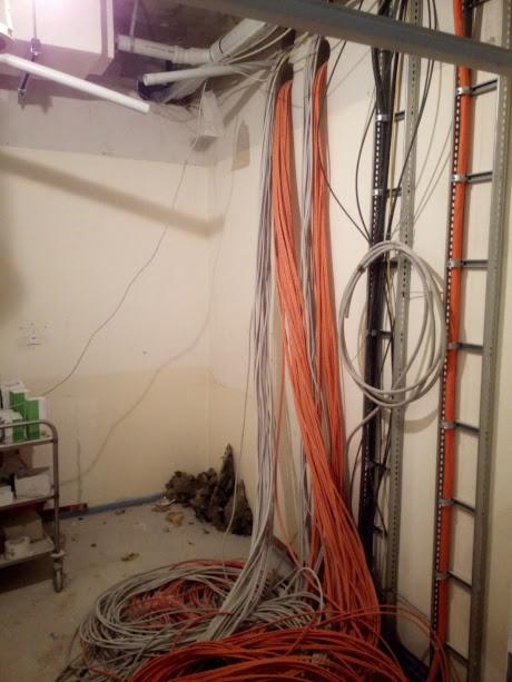 Pomieszczenie w trakcie prac związanych z rozprowadzeniem kabli: wiązki pomarańczowych i szarych przewodów zwisają ze ściany i leżą na podłodze, widoczne metalowe drabinki i korytka kablowe.