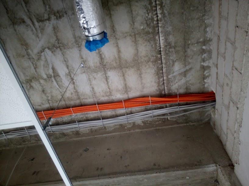 Nierówny betonowy sufit z przymocowanymi wiązkami pomarańczowych i szarych kabli elektrycznych, widoczna instalacja wentylacyjna z niebieskim zabezpieczeniem.