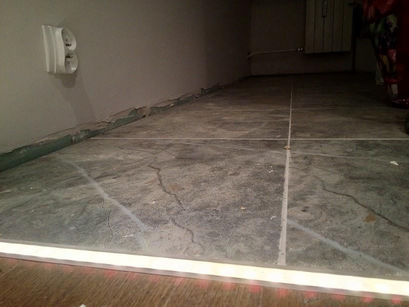 Szare płytki podłogowe z widocznymi fugami, gniazdko elektryczne na ścianie i listwa przypodłogowa z uszkodzeniami.