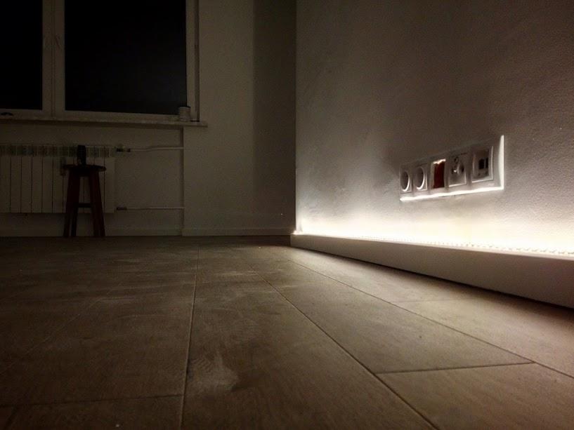 Oświetlenie LED listwy przypodłogowej podkreślające gniazdka elektryczne i włącznik światła na ścianie w nowocześnie urządzonym wnętrzu.