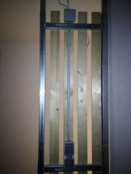 Szara rura instalacyjna przymocowana do drewnianej ściany za pomocą metalowej ramy, z widocznymi puszkami elektrycznymi u góry i dołu.