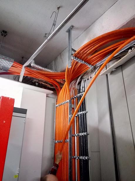 Pomarańczowe i czarne kable prowadzone w metalowych korytach pod sufitem w Radomiu, widoczne mocowania i fragment ręki elektryka.