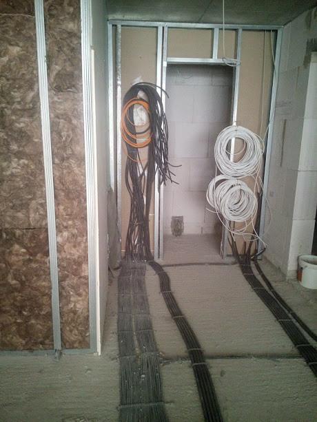 Przewody elektryczne prowadzone wzdłuż ściany z izolacją, widoczne kable czarne i białe w rolkach, oraz pomarańczowe przewody w pętli, w surowym stanie wykończenia wnętrza.