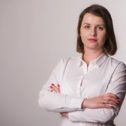 Portret kobiety w białej koszuli z rękami skrzyżowanymi na piersiach, z delikatnym uśmiechem i czerwonymi paznokciami, na jasnym, neutralnym tle.