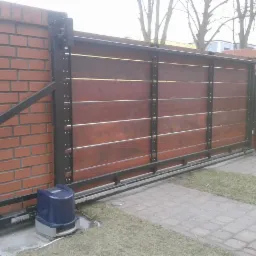 Przesuwna brama wjazdowa z drewnianym wypełnieniem, czarną metalową ramą i napędem elektrycznym, zamontowana na ceglanym murze.