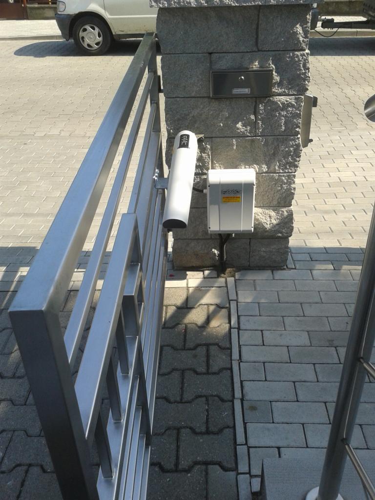 Szara metalowa brama wjazdowa z automatyką, zamontowana na kamiennym słupku z szarą kostką brukową w tle.