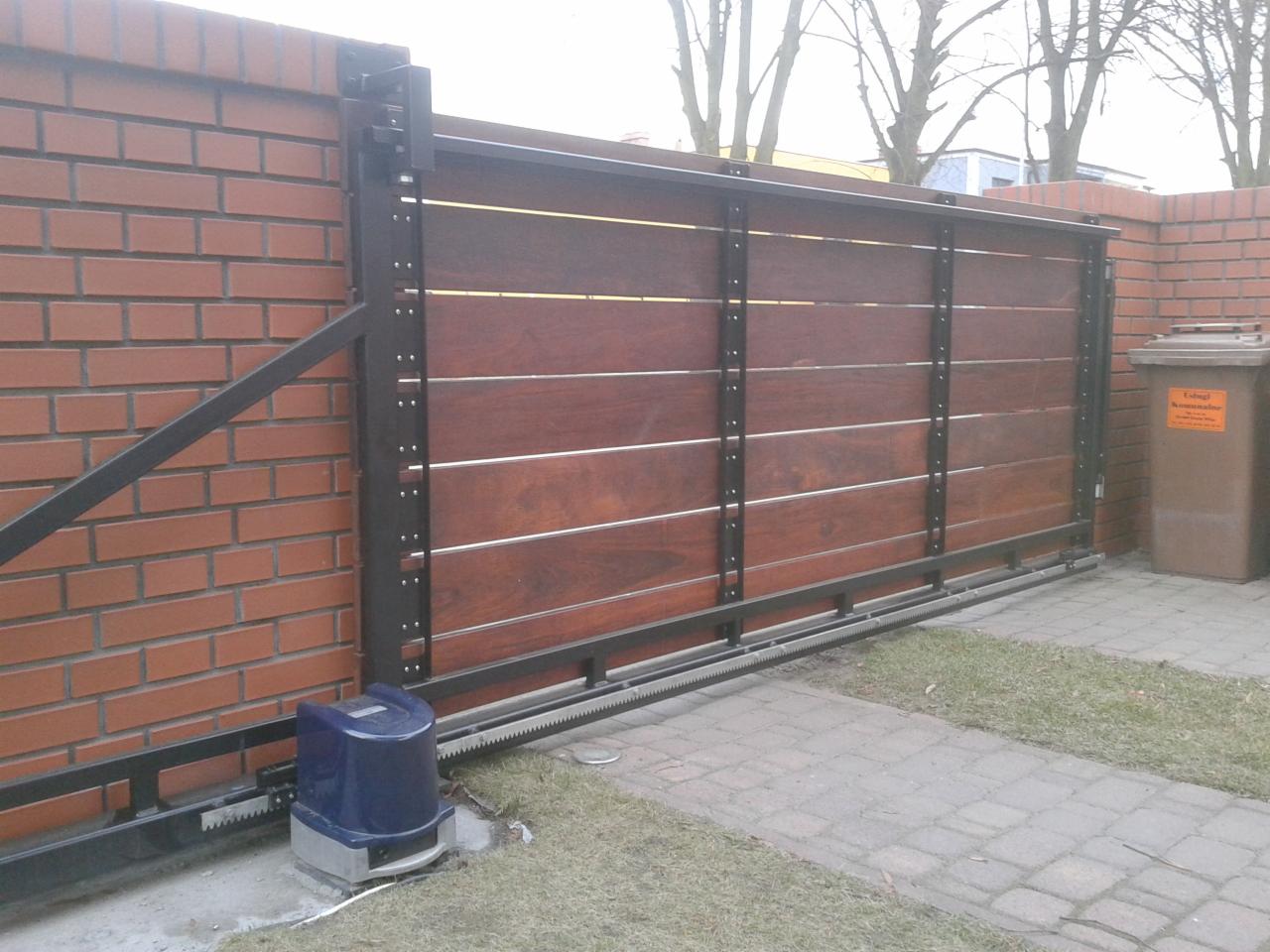 Przesuwna brama wjazdowa z drewnianym wypełnieniem, czarną metalową ramą i napędem elektrycznym, zamontowana na ceglanym murze.