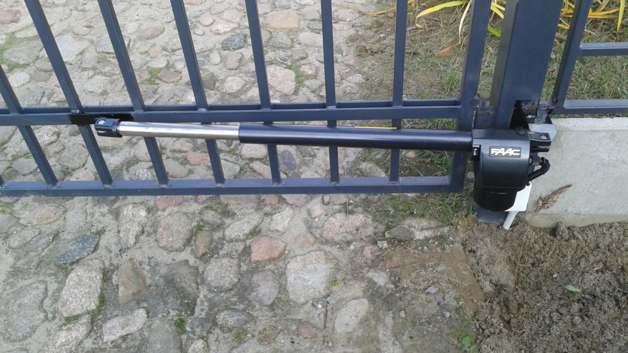 Szczegółowy widok automatyki bramy z siłownikiem FAAC, zamontowanej na metalowej bramie wjazdowej, z brukowanym podjazdem w tle.