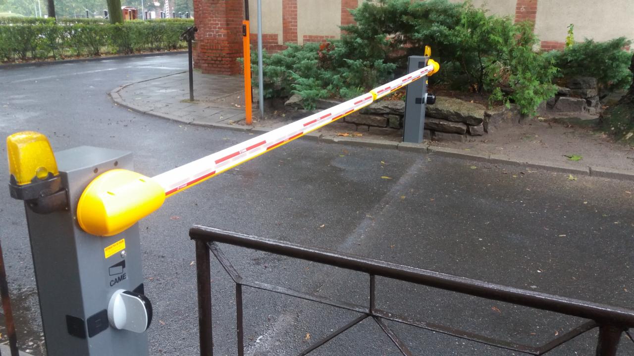 Szlaban automatyczny z żółtą lampą ostrzegawczą, widoczne logo CAME na szarej obudowie, tło z zielenią i mokrą nawierzchnią.