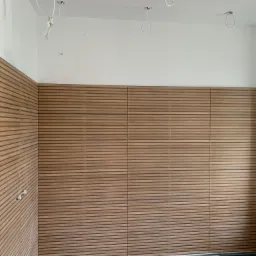 Drewniane panele lamelowe na ścianie w trakcie montażu, widoczne niedokończone kable elektryczne w suficie i fragment okna.