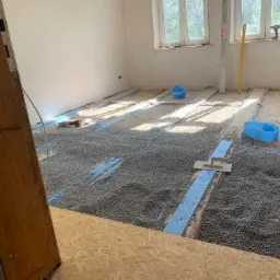 Wnętrze pokoju w trakcie prac przygotowawczych do wylewki, z widoczną warstwą izolacji termicznej z granulatu styropianowego ułożoną między pasami styropianu, narzędzia pracy i fragment sylwetki...