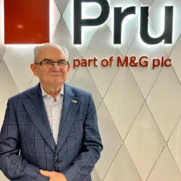 Mężczyzna w okularach i marynarce w kratę stoi na tle podświetlanego logo firmy Pru, będącej częścią M&G plc.