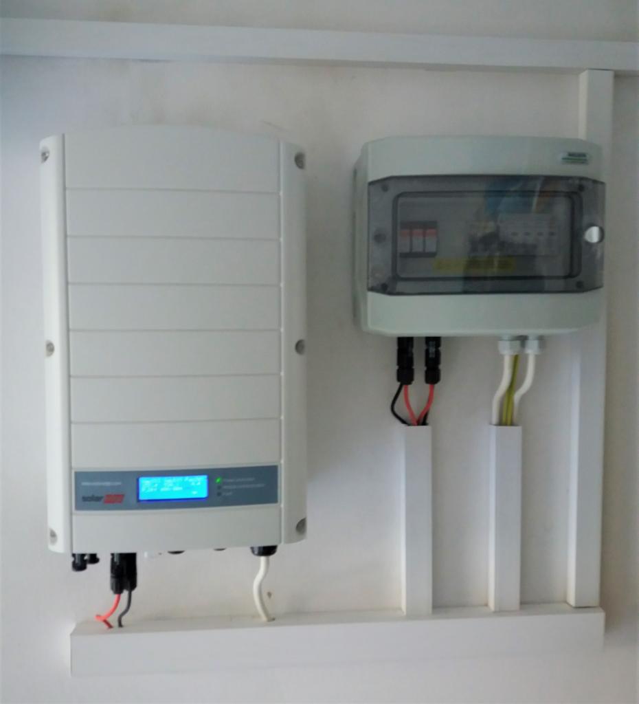 Biały inwerter fotowoltaiczny z wyświetlaczem i skrzynka zabezpieczeń połączone przewodami w instalacji elektrycznej w Radzyniu Podlaskim.