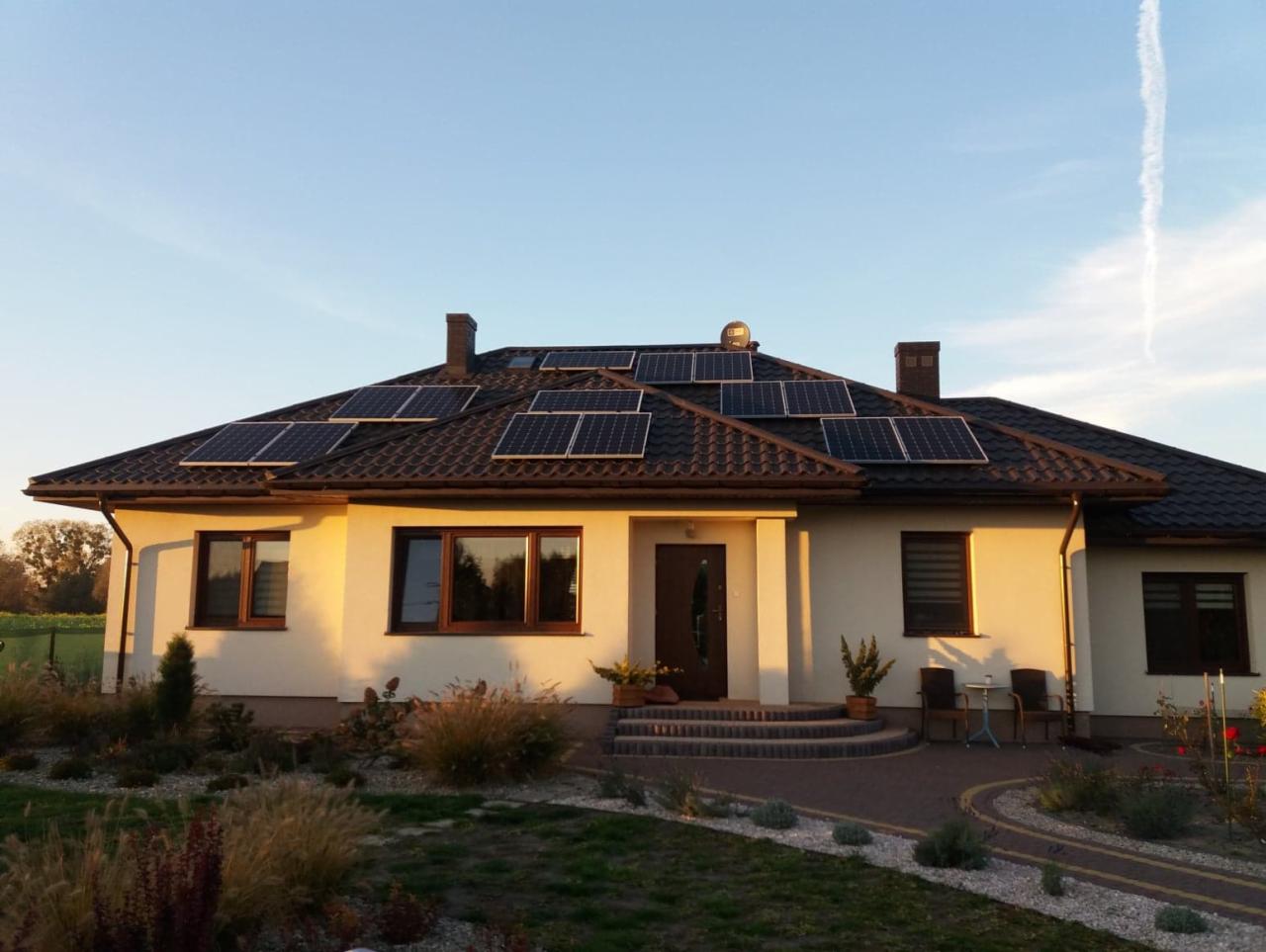 Dom jednorodzinny z brązowym dachem pokrytym panelami słonecznymi; widoczne dwie donice z roślinami przed wejściem, oraz krzesła ogrodowe.