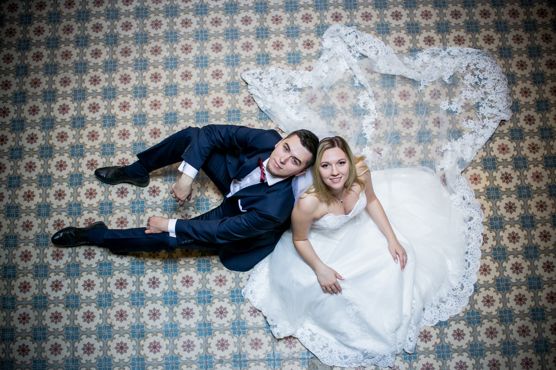 Para młoda leżąca na ozdobnej, wzorzystej podłodze, widok z góry, suknia ślubna z koronkowym welonem rozłożonym obok, granatowy garnitur z bordową muszką.
