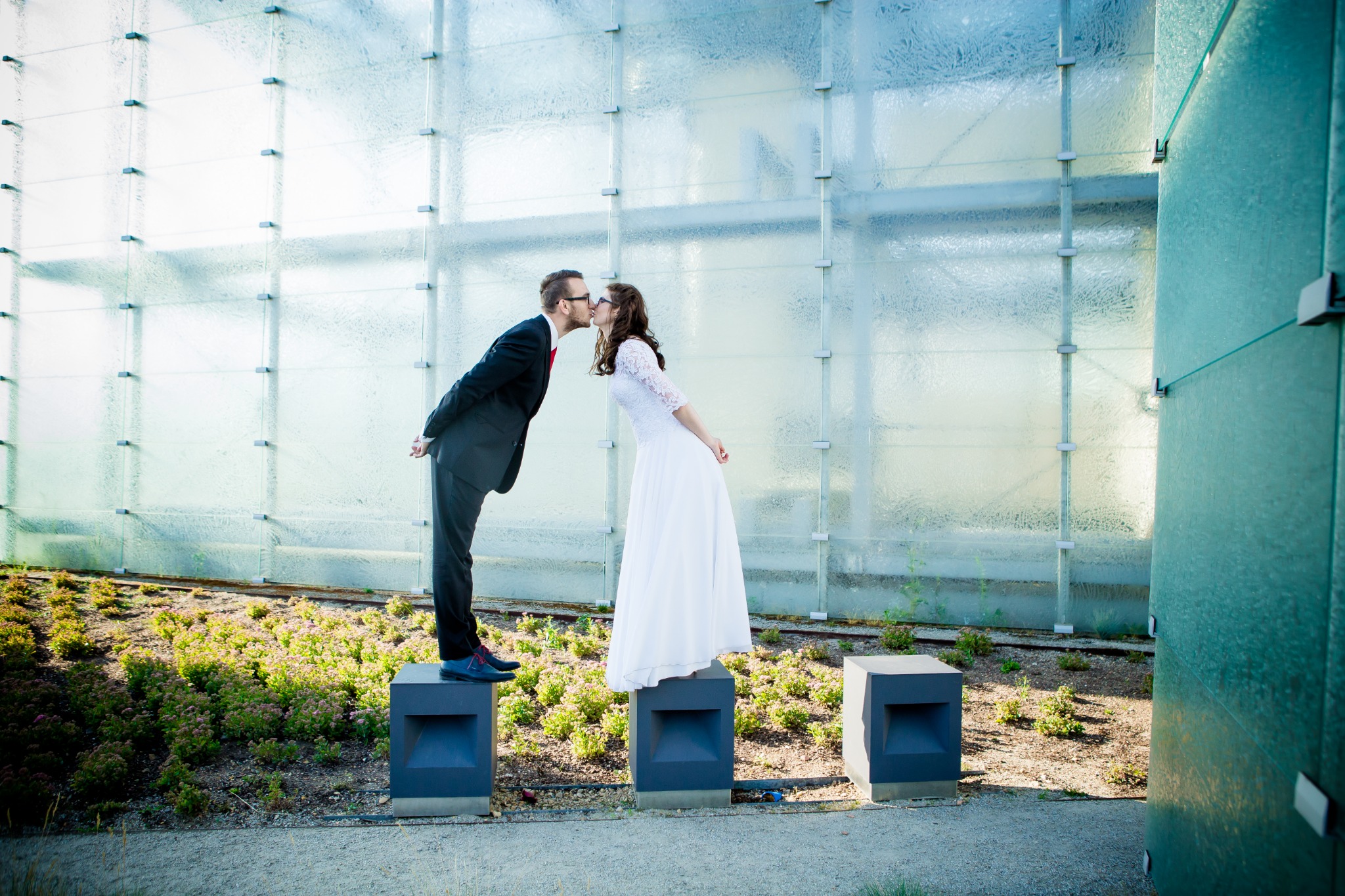 Para młoda w okularach całuje się, stojąc na betonowych postumentach przed budynkiem o szklanej fasadzie.