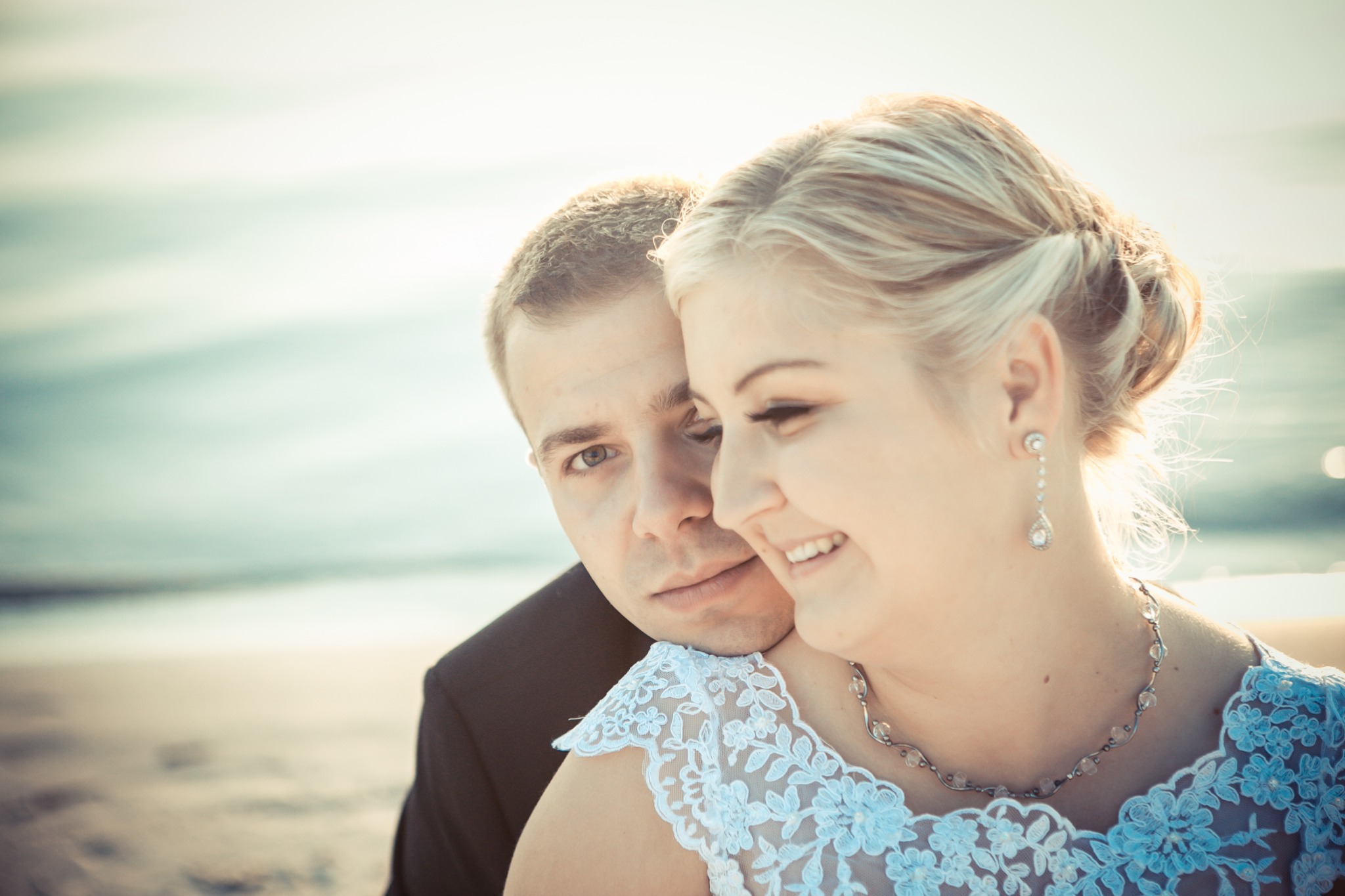 Para młoda na plaży: uśmiechnięta blondynka w koronkowej sukni ślubnej i eleganckim naszyjniku, opierająca się o ramię mężczyzny w czarnym garniturze, z delikatnym morskim tłem.