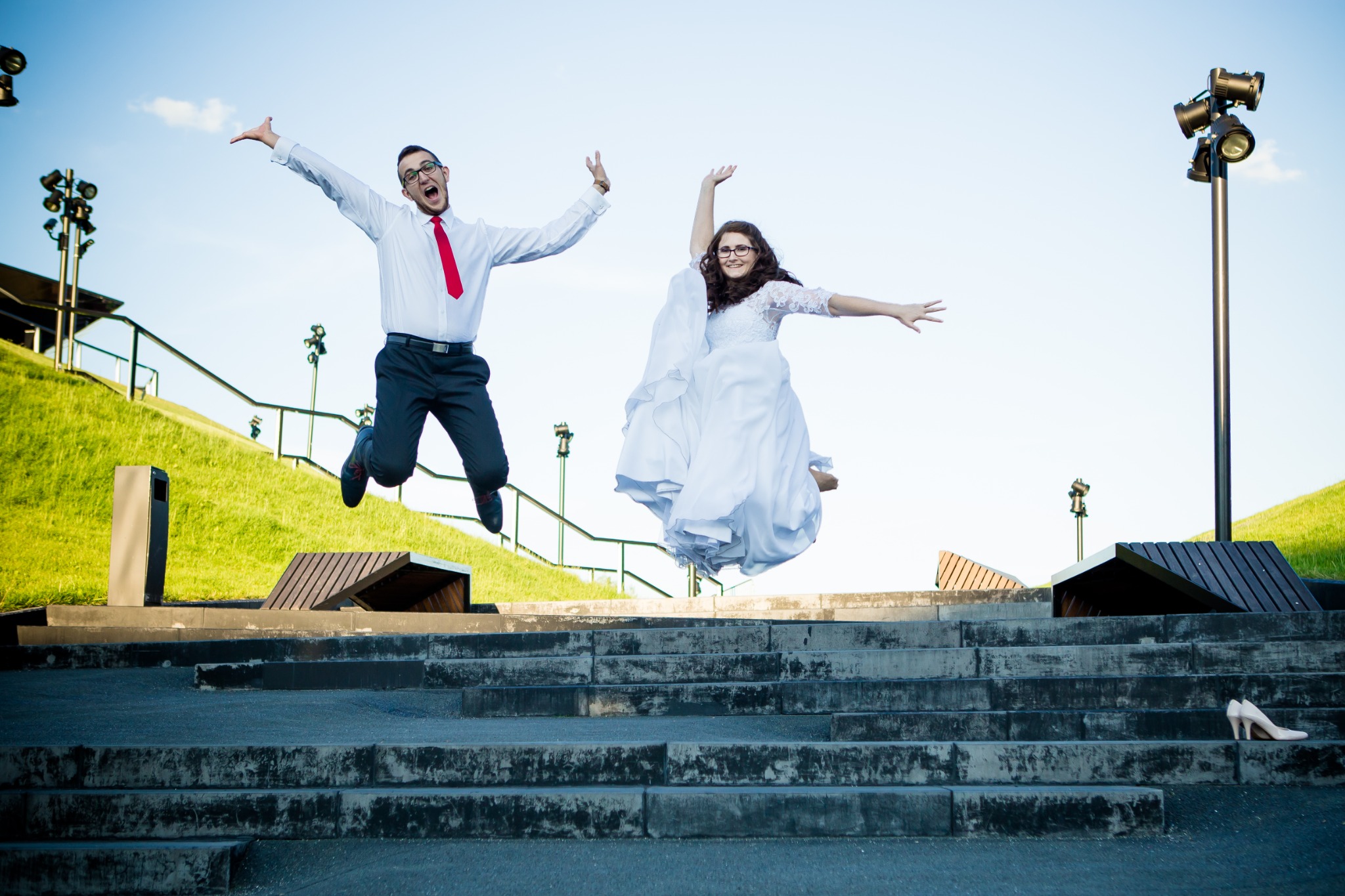 Para młoda w plenerze: Pan młody w skoku z czerwoną muchą, panna młoda w zwiewnej sukni, oboje unoszą się nad betonowymi schodami, na których leżą damskie szpilki.