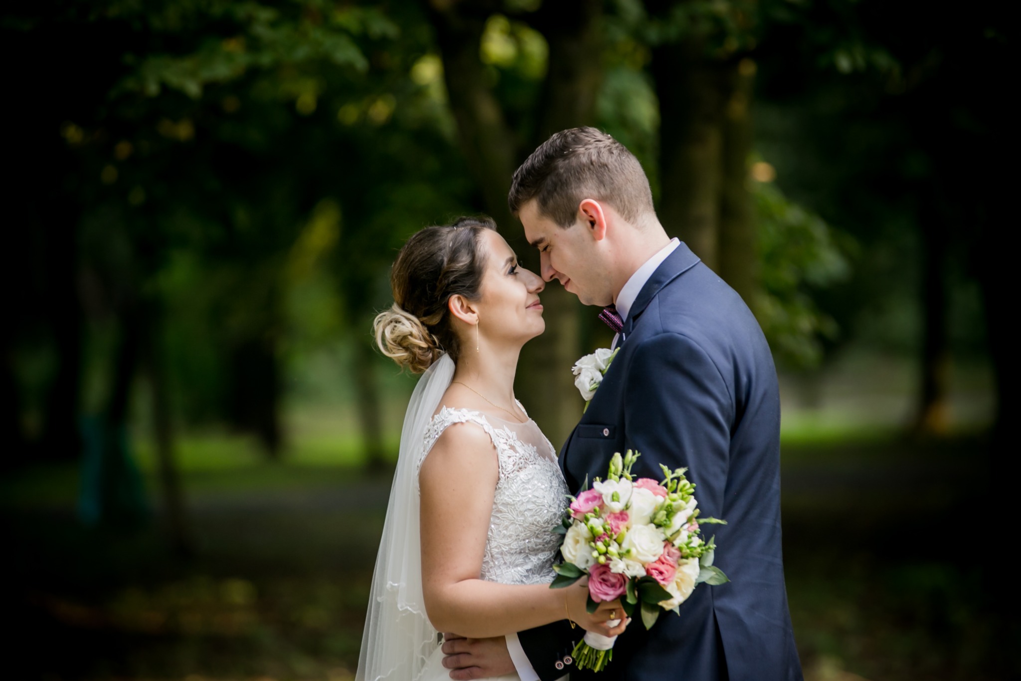 Para młoda, panna młoda w koronkowej sukni i welonie trzyma bukiet różowych i białych kwiatów, pan młody w granatowym garniturze i bordowej muszce, zbliżają się do siebie twarzami na tle zieleni...