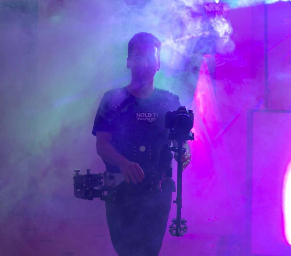 Operator kamery z rigiem i stabilizatorem w zadymionym, kolorowym otoczeniu podczas nagrywania eventu.