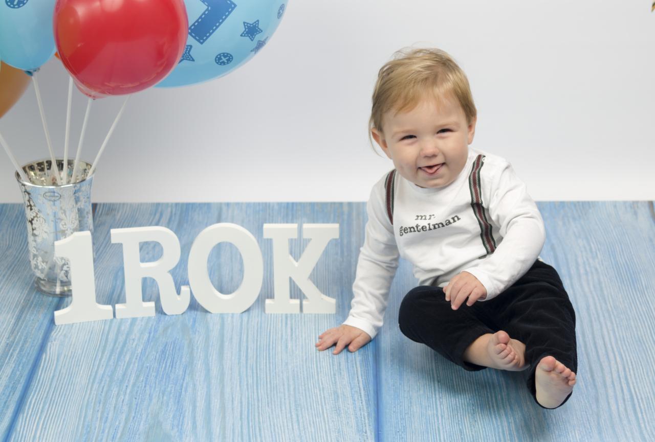 Radosny roczny chłopiec w białej koszulce z szelkami siedzi na niebieskim tle obok balonów i napisu '1 ROK', wystawiając język.