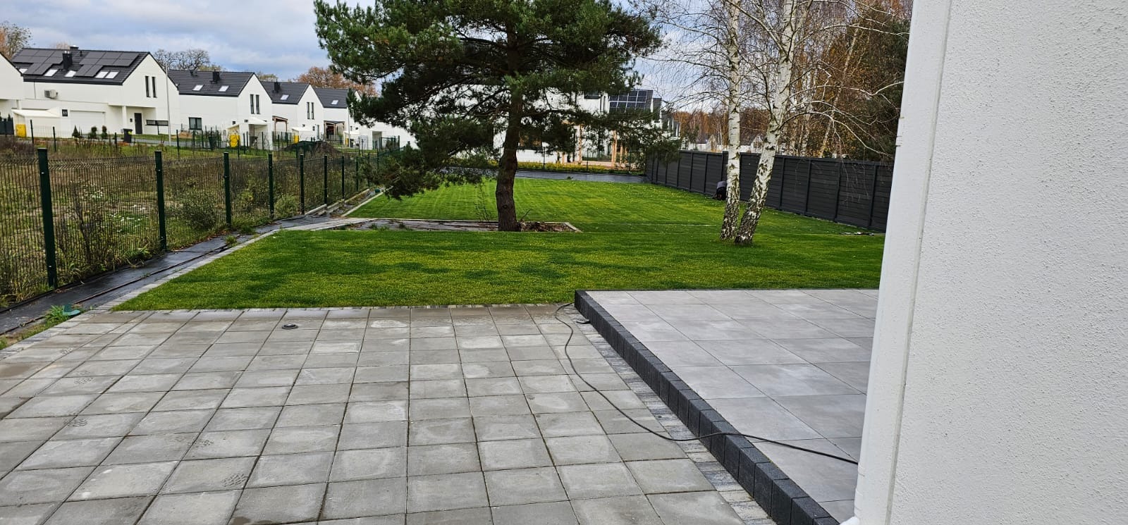 Nowoczesny ogród z szarą kostką brukową, świeżo skoszoną trawą i ogrodzeniem panelowym w tle, widok z rogu tarasu przy białej ścianie budynku.