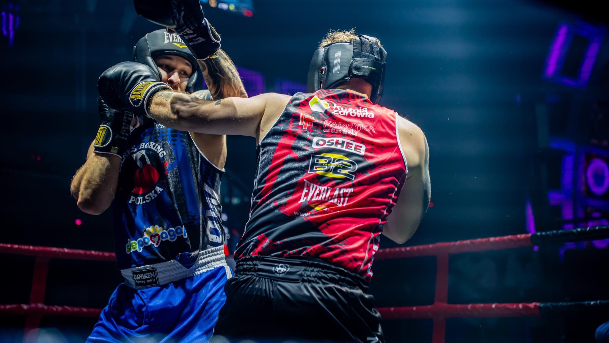 Dwóch bokserów w trakcie walki na ringu, zawodnik w niebieskim stroju z uniesioną rękawicą, zawodnik w czerwonym stroju z logotypami sponsorów na plecach, oświetlenie sceniczne.