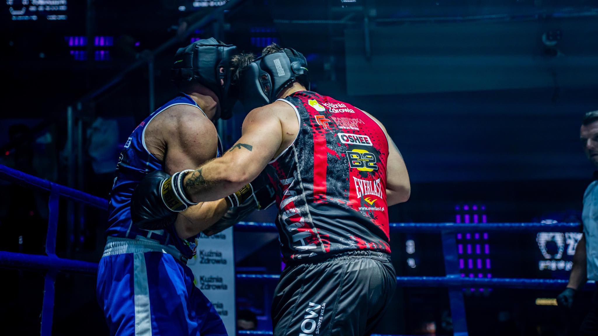 Dwóch zawodników w ringu w trakcie walki, obaj w kaskach i rękawicach bokserskich, jeden w stroju niebieskim, drugi w czerwono-czarnym, widoczne logotypy sponsorów na strojach.