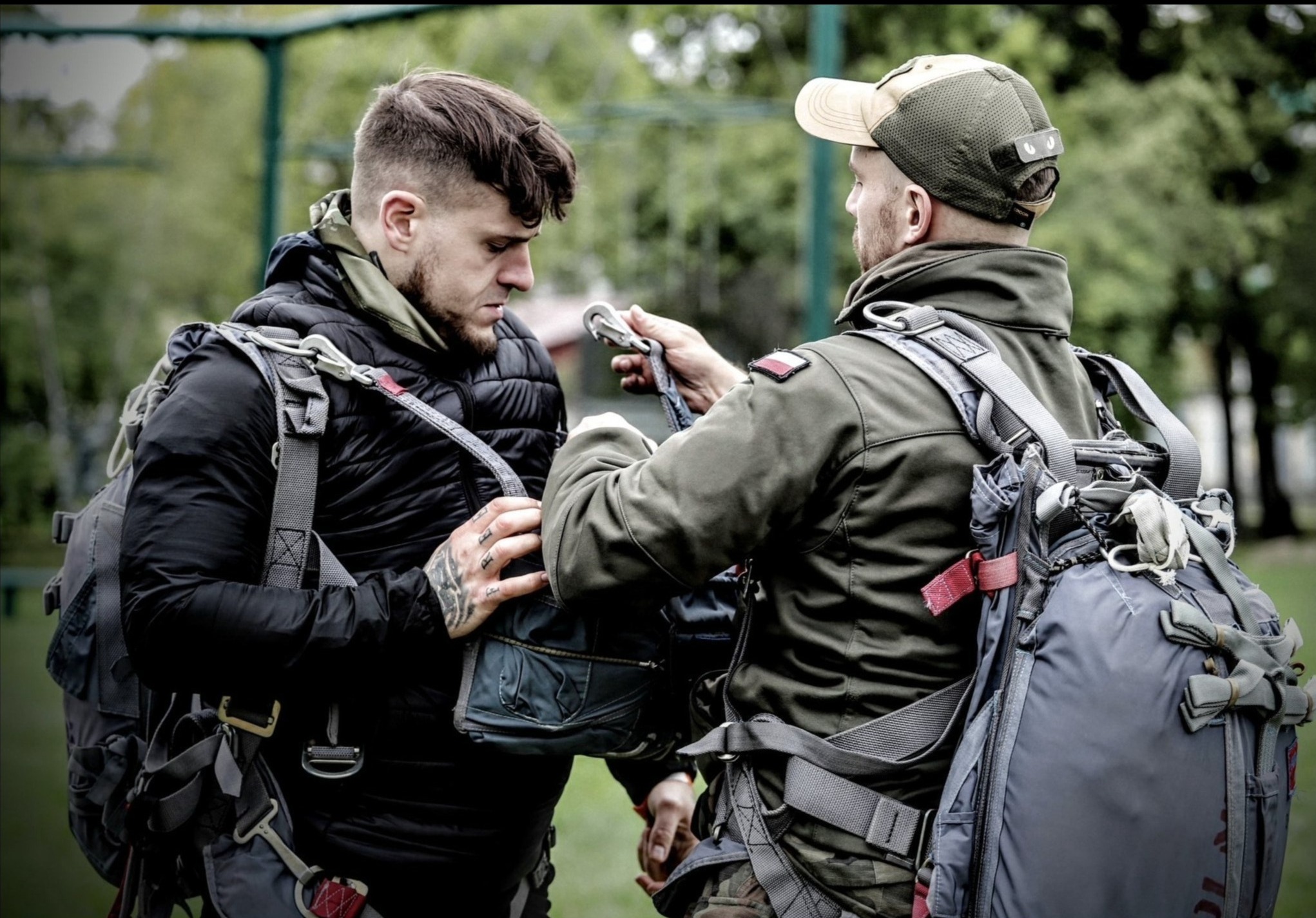 Dwóch mężczyzn w uprzężach spadochronowych sprawdza sprzęt przed skokiem. Jeden z nich ma tatuaże na dłoniach i nosi czarną kurtkę, drugi w wojskowej kurtce z naszywką flagi Polski.