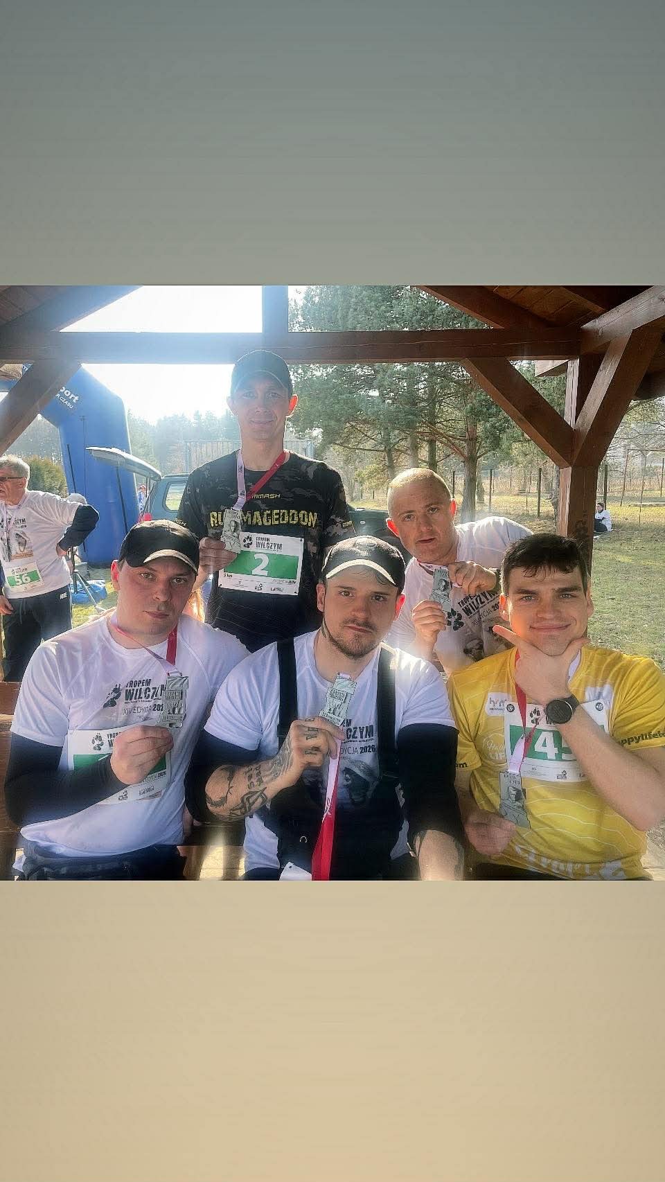 Grupa sześciu mężczyzn pozuje z medalami po biegu, w tle widoczny namiot startowy i las. Uczestnicy w koszulkach z logo biegu i numerami startowymi.