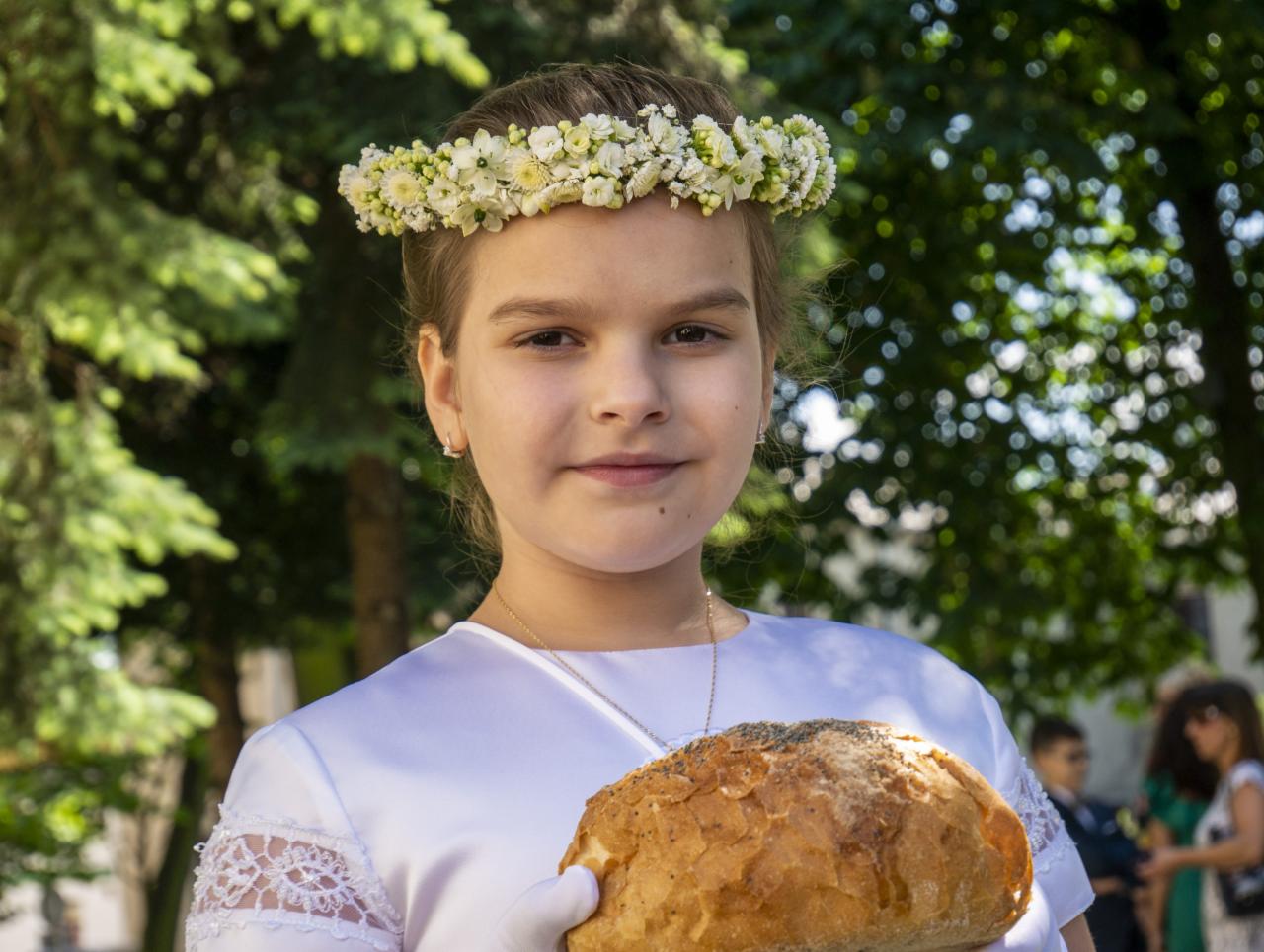 Dziewczynka w białej sukience komunijnej z wiankiem z białych kwiatów na głowie, trzymająca w rękach bochenek chleba z makiem na tle zielonych drzew.