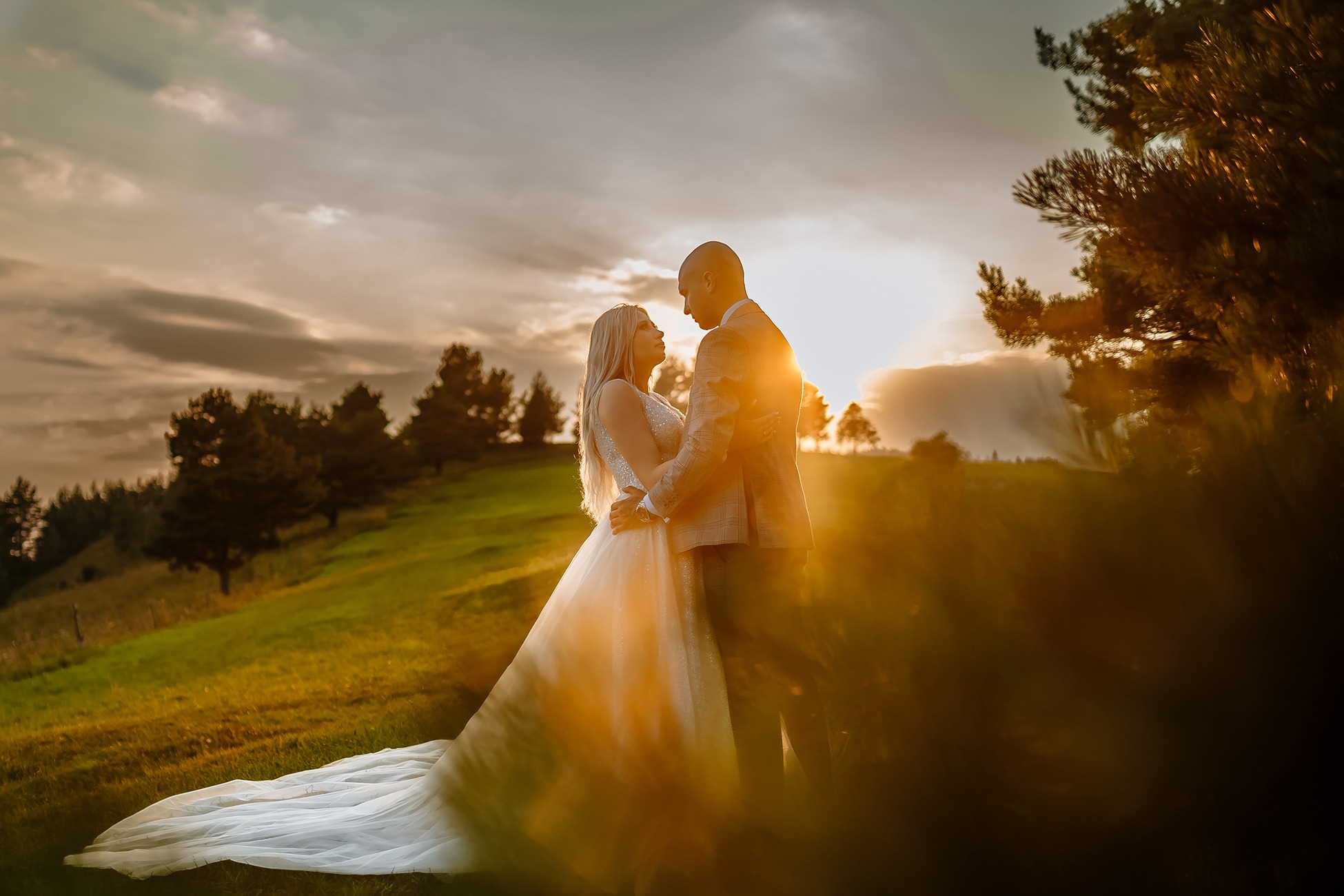 Para młoda w objęciach na wzgórzu o zachodzie słońca, suknia ślubna z długim trenem, garnitur w kratę, ciepłe światło.