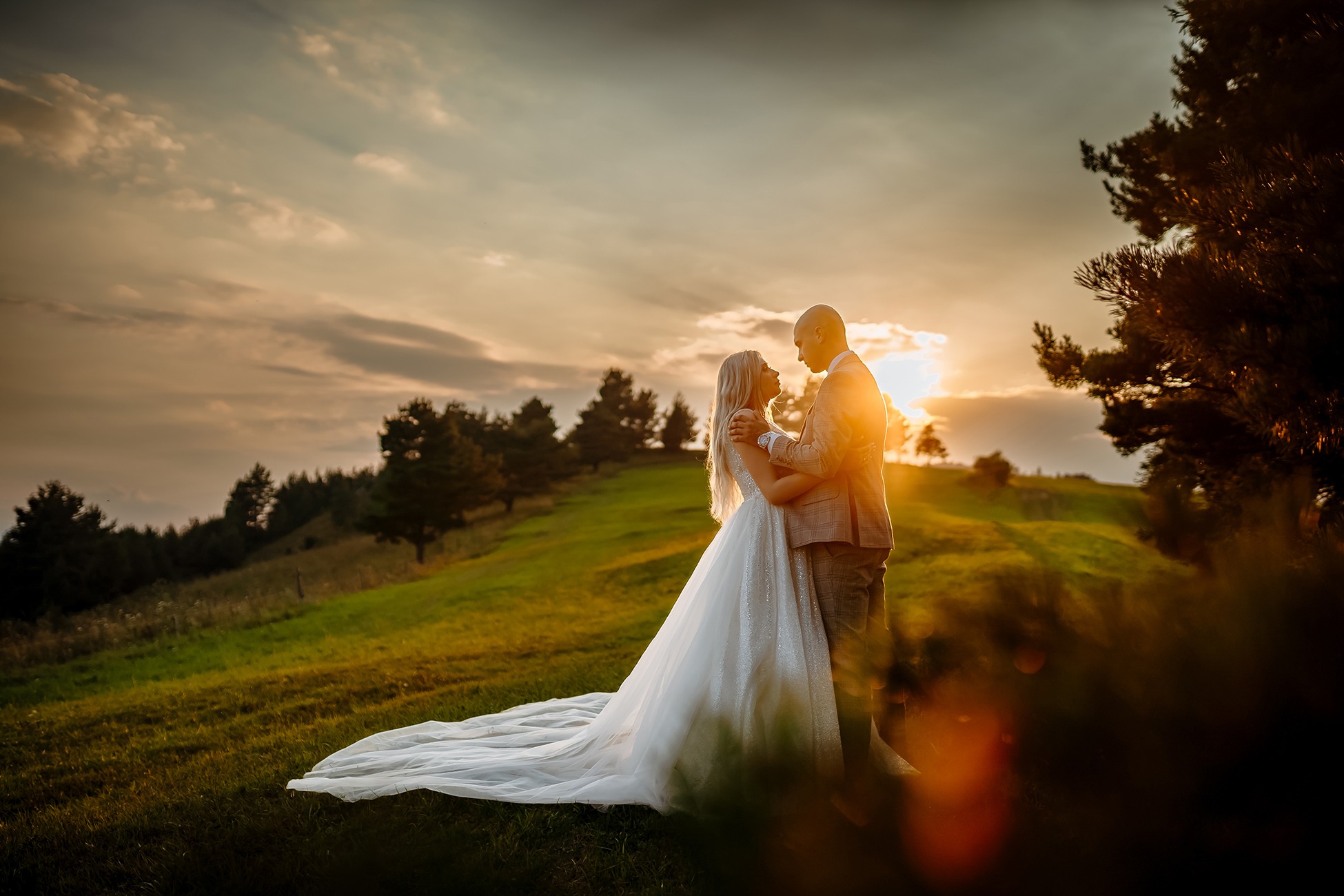 Para młoda w objęciach na zielonym wzgórzu o zachodzie słońca, suknia panny młodej z długim trenem, w tle drzewa i pomarańczowe niebo.