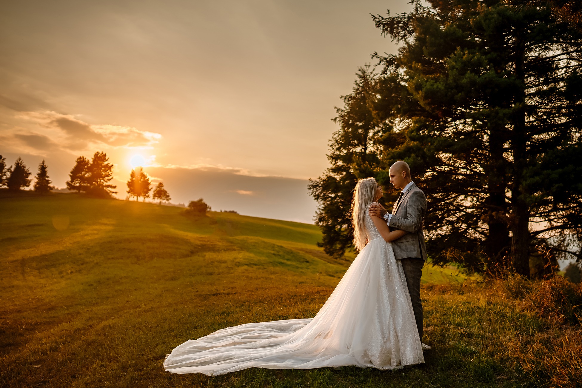 Para młoda w objęciach na wzgórzu o zachodzie słońca, suknia ślubna z długim trenem, pan młody w szarej kracie.