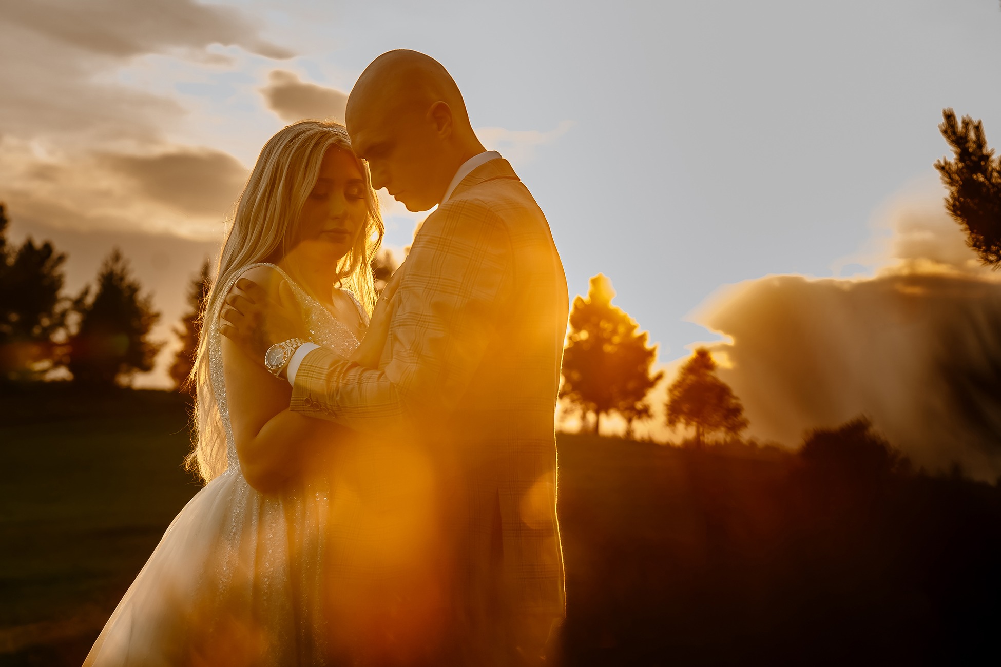 Para młoda obejmująca się w kontraście zachodzącego słońca, panna młoda w sukni z kryształkami, pan młody w beżowym garniturze w kratę.