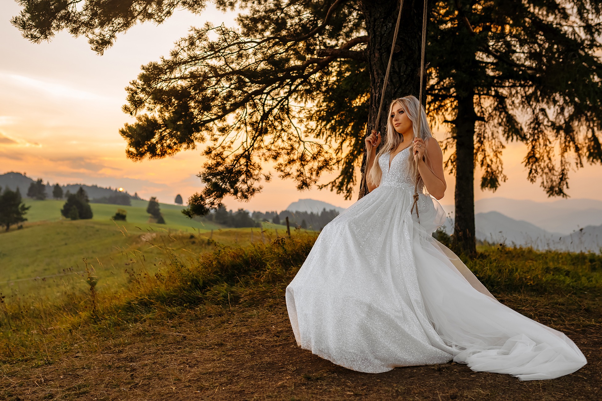 Panna młoda w błyszczącej sukni siedzi na huśtawce zawieszonej na drzewie, w tle zachód słońca nad wzgórzami.
