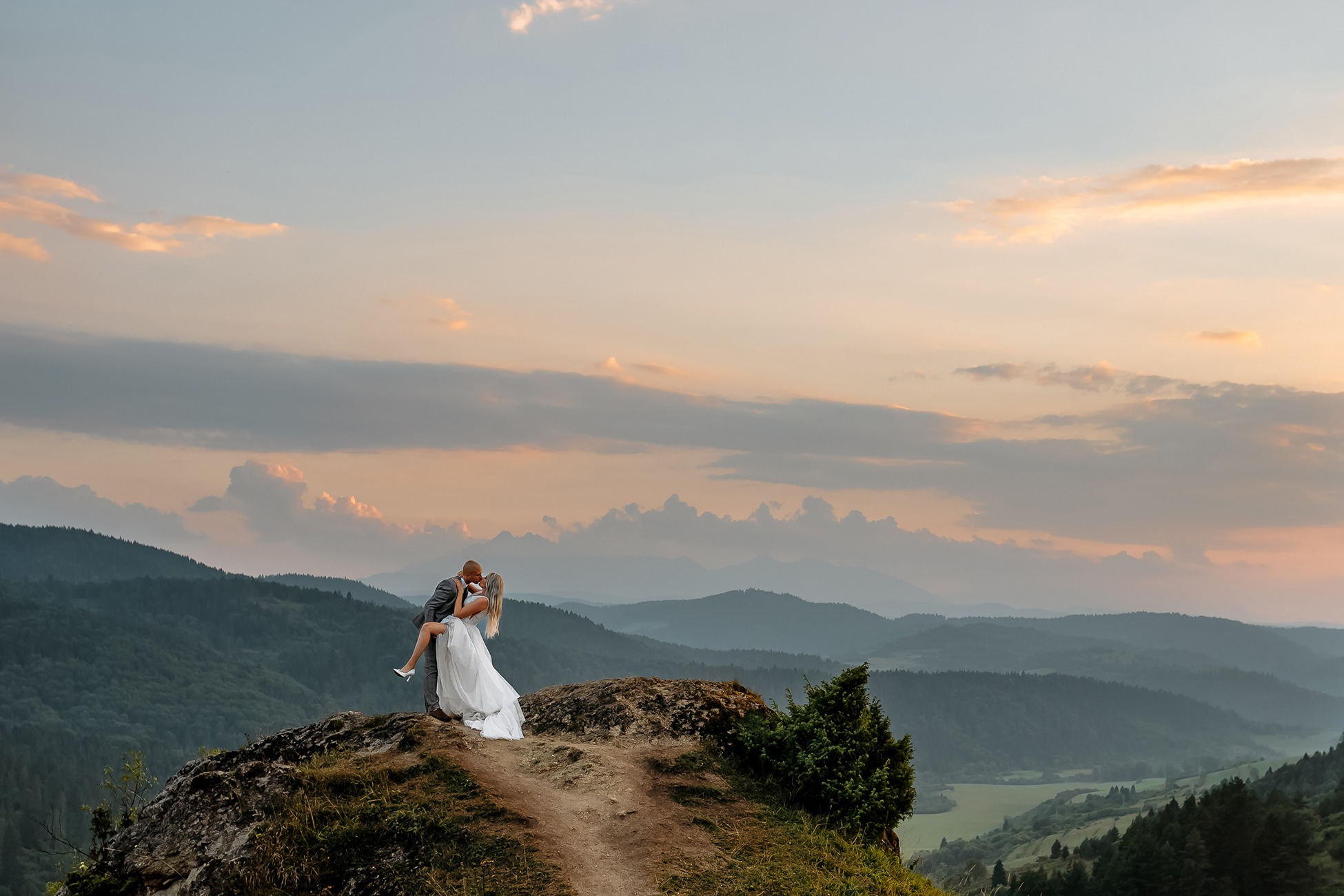 Para młoda w objęciach na szczycie skalistego wzgórza o zachodzie słońca, w tle malownicze góry i niebo z pastelowymi chmurami.
