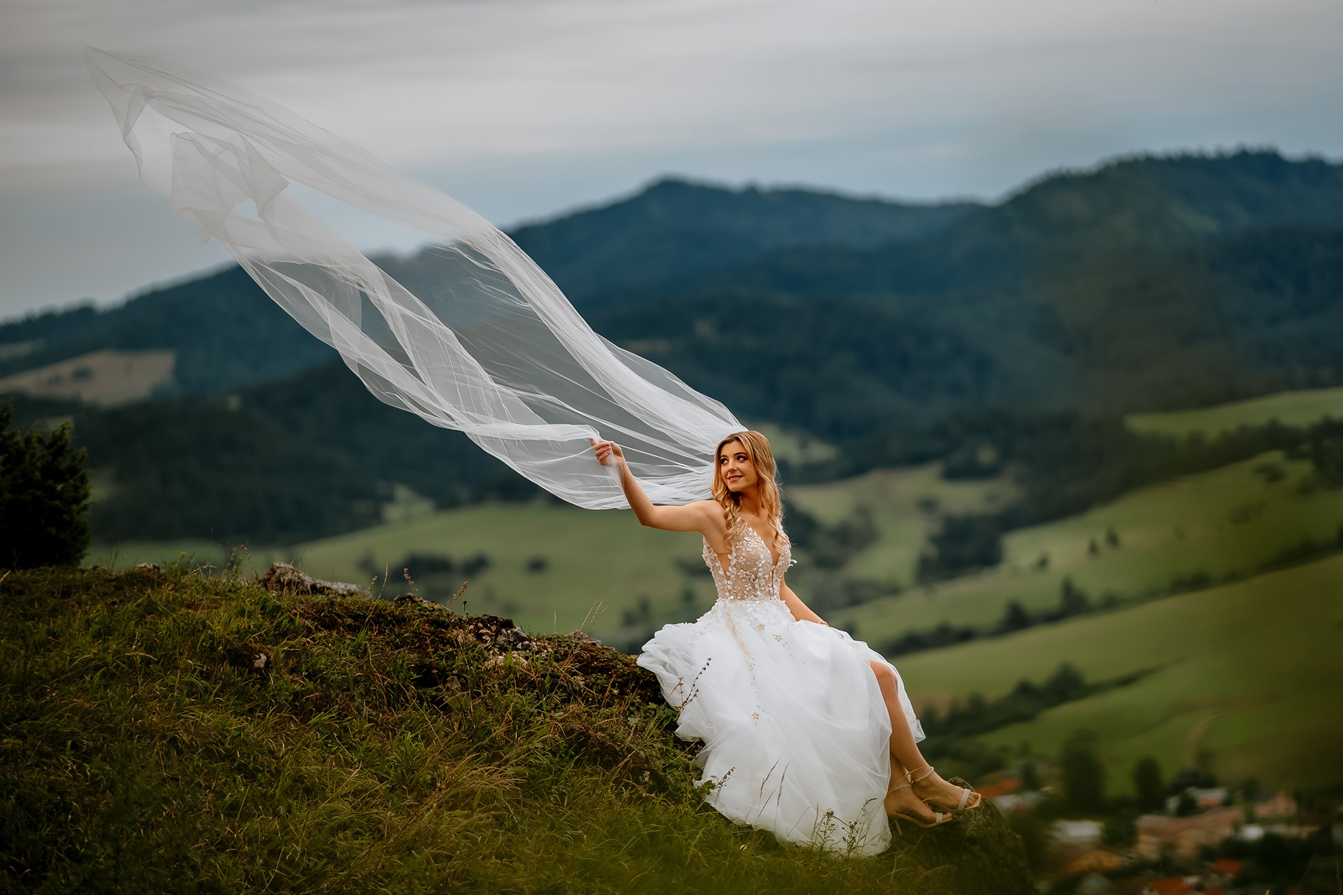 Panna młoda w białej sukni siedzi na wzgórzu w Marcinkowicach, trzymając w dłoni powiewający welon na tle górskiego krajobrazu.